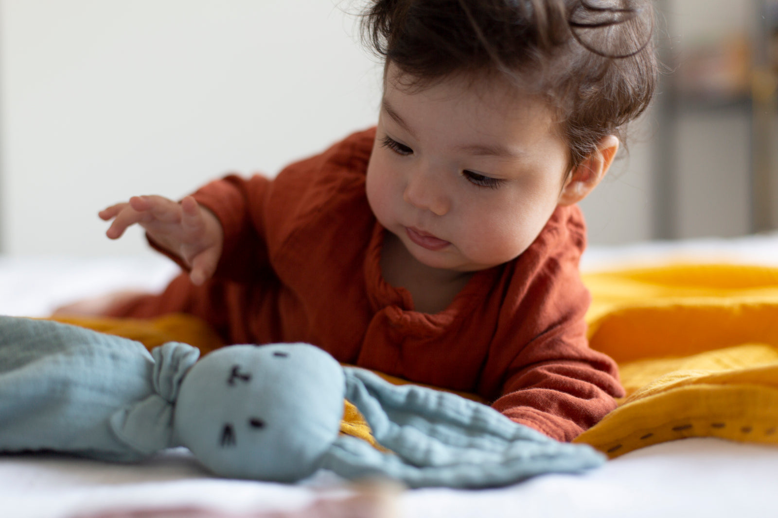 Boy with rust baby suite on mustard playing cloth is reaching for sea green bunny.